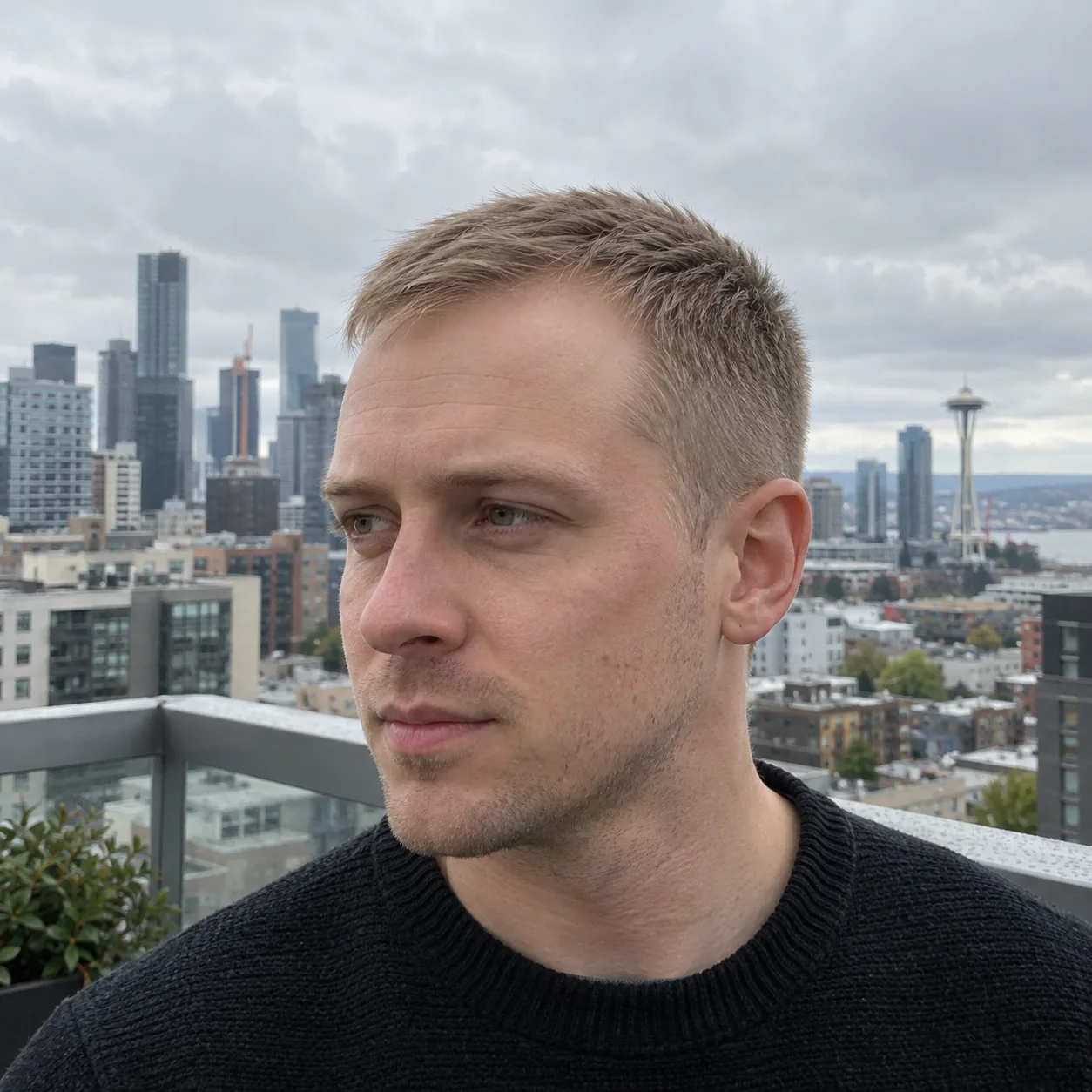 Young man with fine ash blonde hair cut in a short blunt crop worn forward, on a balcony above a city skyline