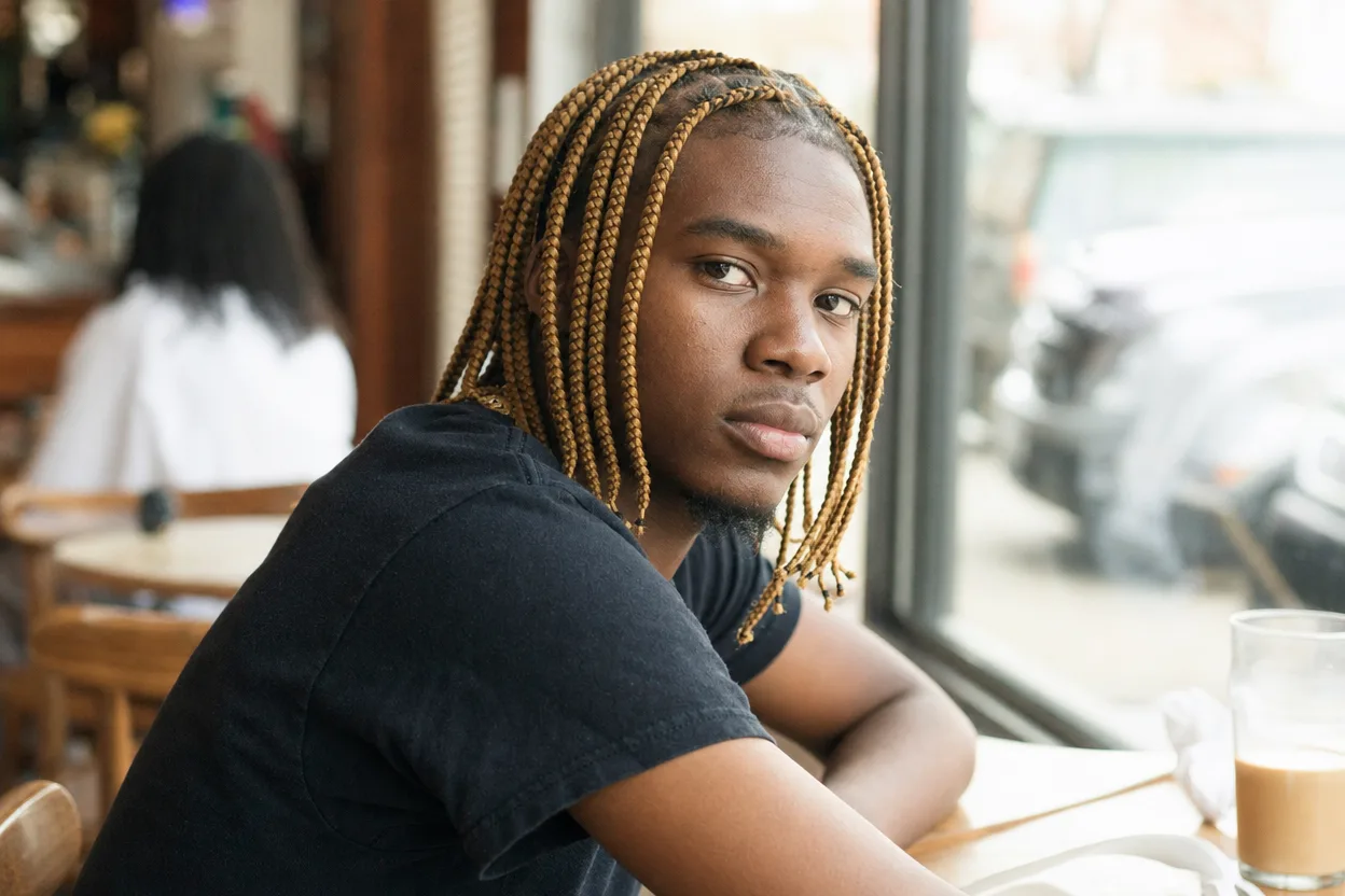 Young man with honey blonde box braids hanging to his shoulders, seated at a cafe table by a window