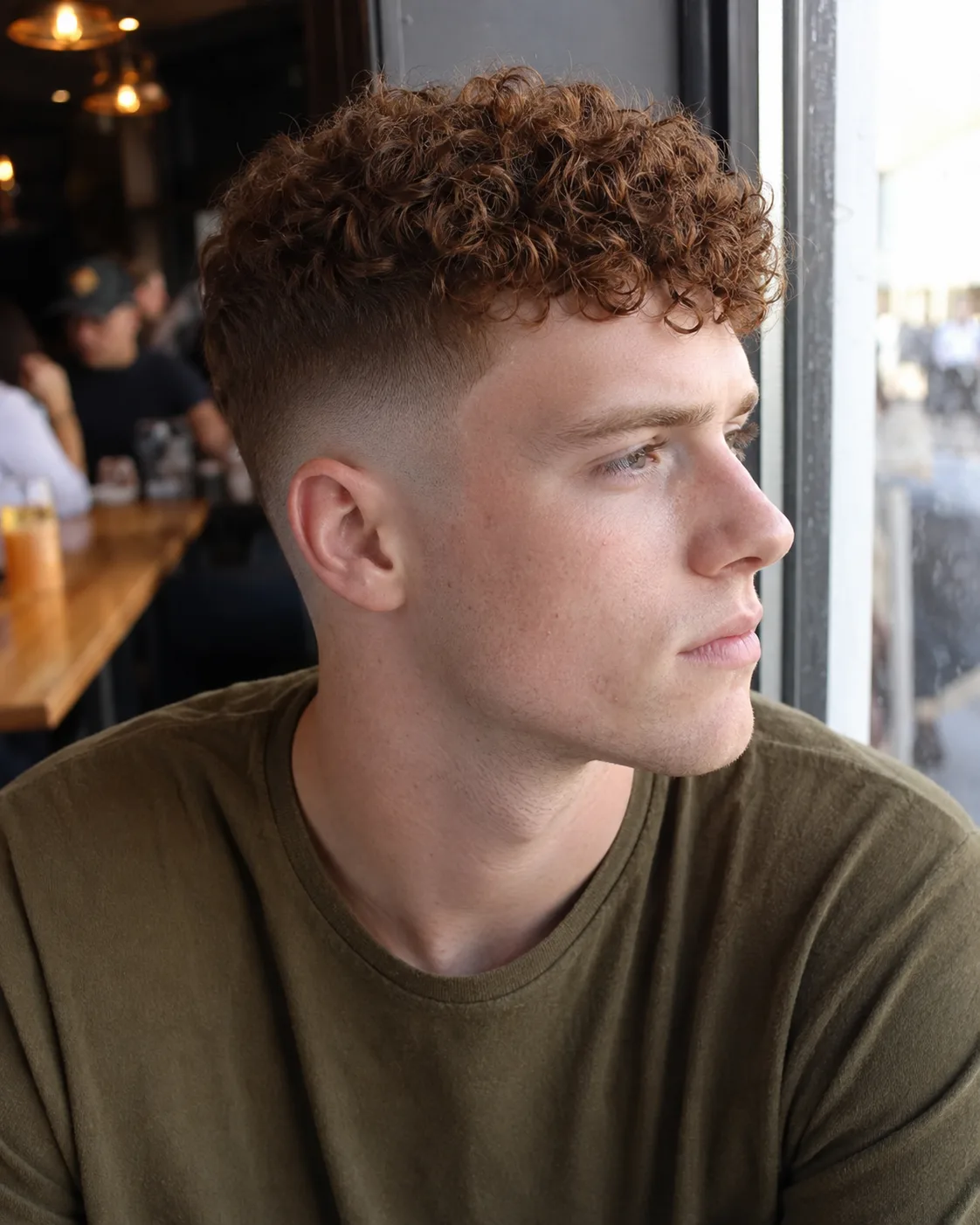Young man with long auburn curls on top sitting by a cafe window, the entire side of his head clipped bare from the sideburn up past his ear